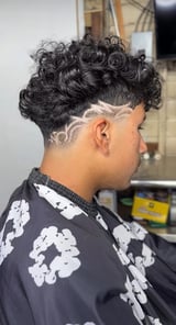 Side profile of a person with a curly black textured hairstyle, undercut with geometric line design, wearing a black patterned shirt