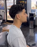 Man with styled black hair and facial hair wearing a gray shirt, seated in a barber shop interior
