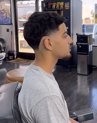 Man with styled black hair and facial hair wearing a gray shirt, seated in a barber shop interior