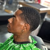 Man in profile wearing green patterned shirt with short braided hairstyle and ear piercing in barber shop setting