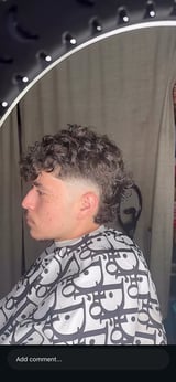 Man with curly hair fade haircut wearing patterned black and white shirt in front of vanity mirror with lights