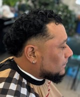 Side profile of a man with a textured curly fade hairstyle, facial hair, and wearing a checkered shirt