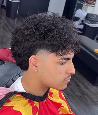 Young man with curly dark hair and a modern undercut hairstyle, wearing a red and yellow striped shirt, photographed in profile inside a barbershop
