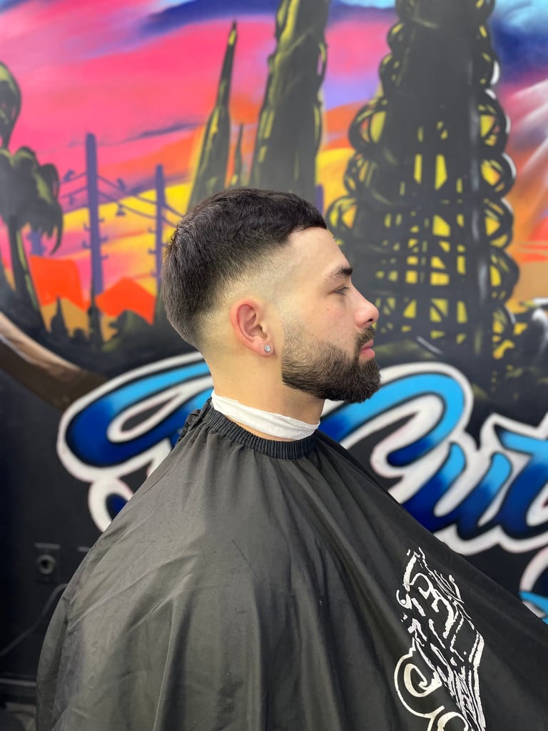Man with a close-cropped fade haircut and beard in profile, wearing a black barber cape, in front of a colorful cityscape mural
