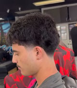 Side profile of a person with short dark textured hair in a barbershop setting, wearing a red and black plaid shirt