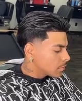 Man with styled dark hair undercut and gold hoop earring, wearing patterned black and white shirt in barbershop setting