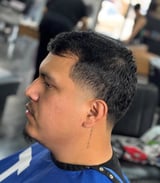 Man with dark hair and goatee in profile view, wearing blue and black shirt, inside a barbershop