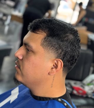 Man with dark hair and goatee in profile view, wearing blue and black shirt, inside a barbershop
