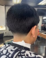 Back view of a person with short black hair and a fade haircut, wearing a patterned dark and white shirt in a salon setting